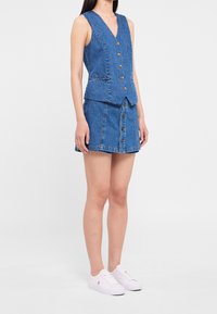 Denim väst och kjolset i ljusblått. Vesten har V-hals, knappar fram och två fickor. Kjolen har knappstängning med fransar i nederkant.