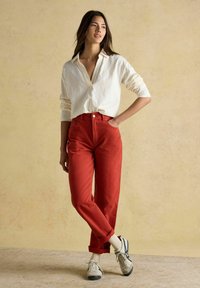 Naisel on beezil seinal seismas, seljas valge pika varrukaga särk, kõrge vöökohtadega punased püksid, valged sokid ja beežid tennisetsid, millel on tumesinised detailid.