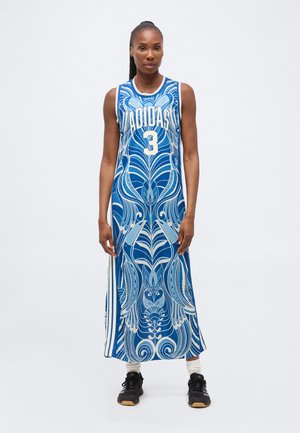 Femeie în picioare, purtând o rochie lungă albastră fără mâneci Adidas, cu modele abstracte albe, șosete albe și adidași negri pe un fundal simplu.