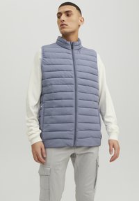 Light gray puffer vest with horizontal quilted design, zipper front, high collar, and a smooth, matte texture. Worn over a white long-sleeve shirt.