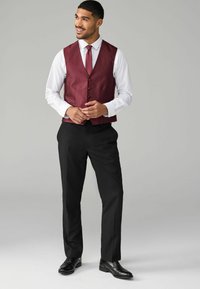 Bordový vestový kabát s piatimi gombíkmi, skombinovaný s bielou košeľou a čiernymi nohavicami. Čierne formálne topánky dotvárajú celý outfit. Celkový jemný vzhľad.