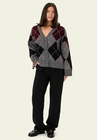 Grauer Argyle-Strickjacke mit burgunderroten, schwarzen und weißen Rautenmuster, mit tiefem V-Ausschnitt und großen Knöpfen vorne, kombiniert mit schwarzen Hosen.