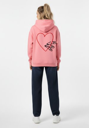 Felpa rosa con un grande design a cuore rosso e spilli di sicurezza neri sul retro, abbinata a pantaloni blu scuro e sneakers bianche.
