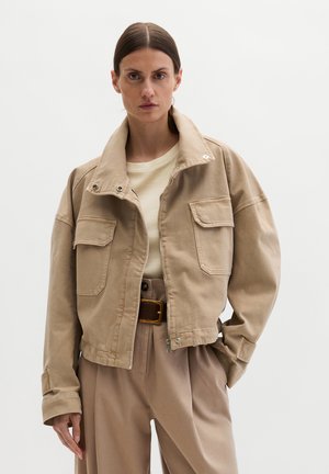 Frau trägt eine beige kurze Jacke mit Klapptaschen über einem hellen Hemd, hoch taillierte beige Hose und einen braunen Gürtel.