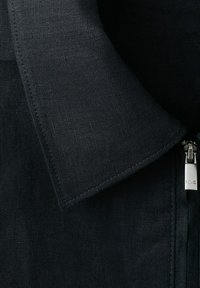 Zwarte gestructureerde jaskraag met zichtbare stiksels en een zilveren ritstrekker aan de rechterkant, gegraveerd met "MNG".