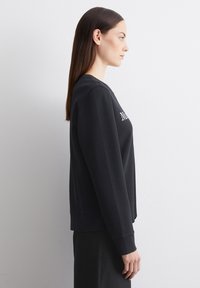 Musthave mustka, millel on vabalt kukkuv lõige, ümar kaelus ja pikad varrukad. Esiosas on valge tikandiga tekst. Silmapaistev tekstuur ja minimalistlik disain.