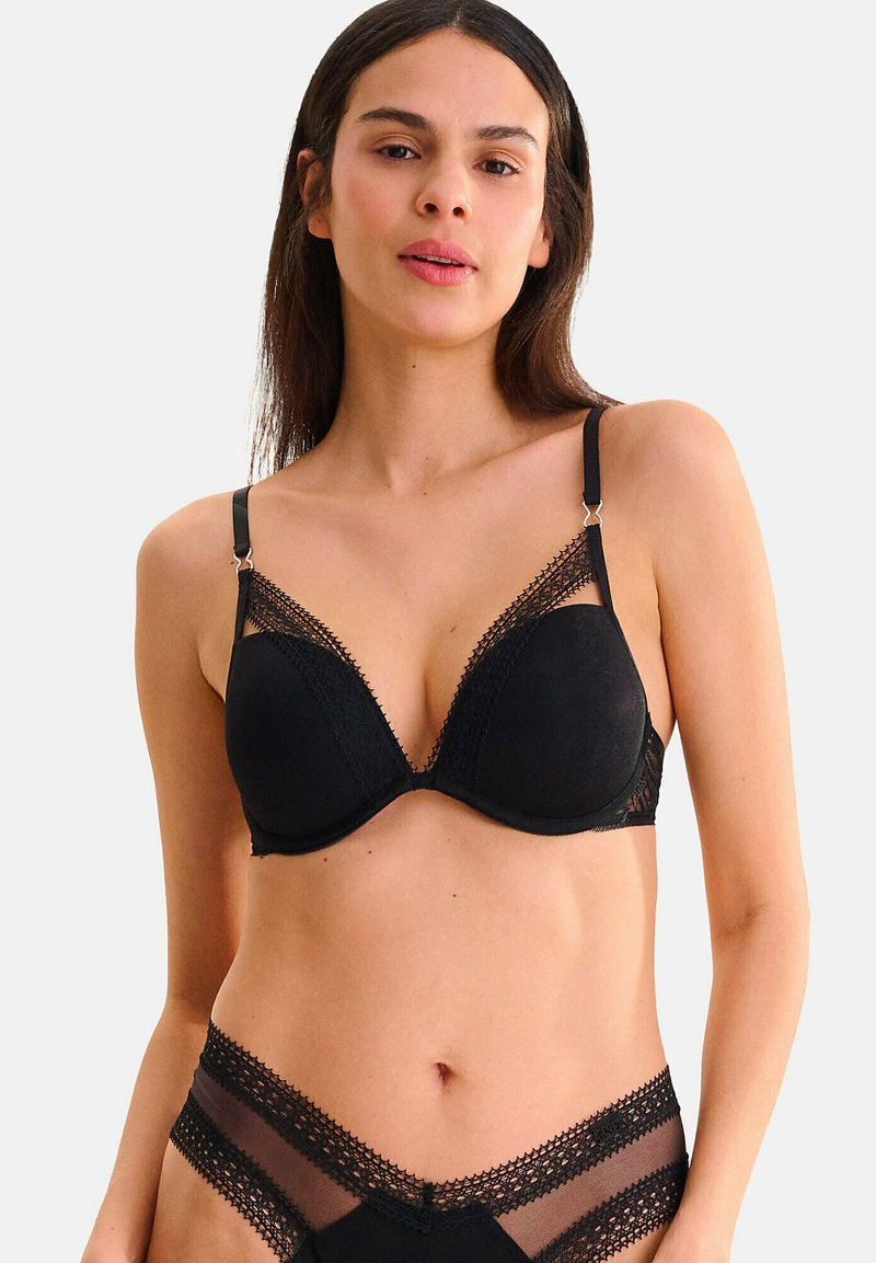 Femme portant un soutien-gorge noir en dentelle avec des bordures et une culotte haute en résille assortie, se tenant contre un fond clair uni.