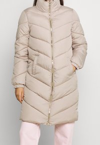 Abrigo acolchado beige con cuello alto, diseño de rombos, cremallera dorada y puños elásticos. Se pueden ver dos bolsillos laterales.