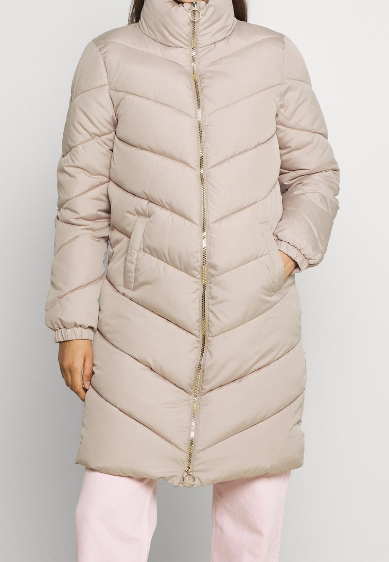 Abrigo acolchado beige con cuello alto, diseño de rombos, cremallera dorada y puños elásticos. Se pueden ver dos bolsillos laterales.