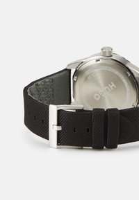 Correa de reloj de tela negra texturizada con una hebilla metálica plateada, que presenta una caja de acero inoxidable y un fondo resistente al agua.