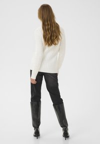 Maglione bianco a coste con maniche lunghe, abbinato a jeans alti scuri e stivali neri con tacco fino al ginocchio. Sfondo neutro.