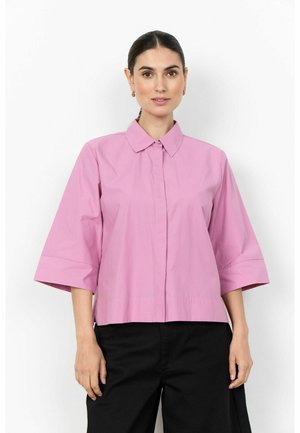 Femme portant un chemisier rose à manches courtes avec col et un pantalon noir, debout devant un fond blanc uni.