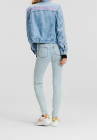 Osoba v světle modré džínsové bundě se nápisem "KARL LAGERFELD" a odpovídajících slim džínách, doplněná bílými teniskami s barevnými logy.