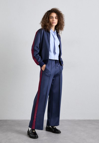 Chándal de color azul marino que incluye una chaqueta corta con franjas rojas, combinada con pantalones de pierna ancha. Los mocasines negros y una camisa de color azul claro con cuello completan el look.