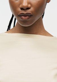 Κοντινό πλάνο ενός ατόμου με σκούρο δέρμα και πλεγμένα μαλλιά που φοράει ανοιχτό μπεζ πουκάμισο με στρογγυλή λαιμόκοψη.