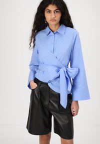 Chemisier bleu clair à manches cloche longues et lien à la taille, associé à un short en cuir noir avec poches latérales.