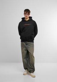 Svart hettegenser med lommer foran og oransje tekstdesign, kombinert med løse denimjeans og beige joggesko.