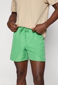 Costume de baie verde, cu talie elastică și șnur, având o textură fină și un branding subtil pe marginea inferioară.