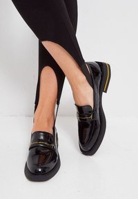 Mocassins en cuir verni noir avec un bout rond, ornés de coutures jaunes et d'une semelle noire épaisse. Portés avec des leggings noirs.