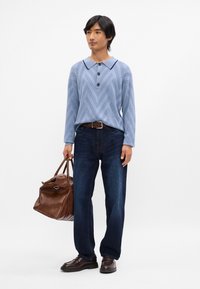 Chemise à manches longues bleu clair à motif chevron avec trois boutons, jeans en denim foncé, sac en cuir marron et chaussures foncées.