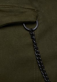 Oliijfkleurige stof met een klapzak, voorzien van een zwart metalen ring- en kettingdetail, wat zorgt voor een textuurrijk contrast.