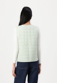 Grön gingham-quiltad väst med en design på baksidan, rund hals och ärmlös stil, i kombination med en vit långärmad topp och blå jeans.
