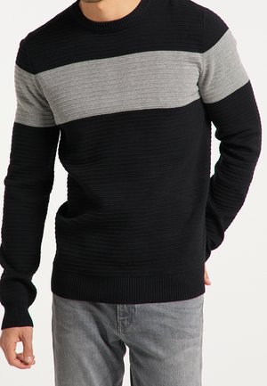 Homme portant un pull en maille côtelée avec une large bande horizontale grise sur la poitrine et du noir sur les manches et le bas du torse.