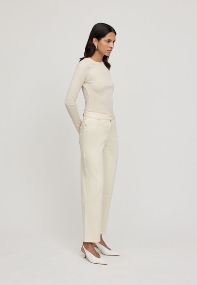 Maglietta a maniche lunghe color crema, pantaloni a vita alta beige a gamba larga e scarpe bianche con punta affusolata. Texture liscia e aderente con cuciture e rifiniture sottili.