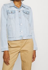 Světle modrá džínová bunda s klasickým designem, obsahující kovové knoflíky, náprsní kapsy a opotřebované detaily. Dlouhé rukávy s manžetami.