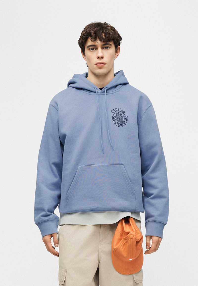 Joven con sudadera con capucha azul claro con detalle de texto en espiral, pantalones cargo beige, sosteniendo una gorra naranja frente a un fondo simple.