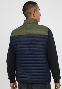 Paminkštintas liemuo, turintis žalią viršutinę dalį ir tamsiai mėlyną apatinę dalį su horizontaliais siūlėmis. Aukštas kaklas ir jokie matomi įrenginių detalės.