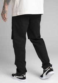 Person som bär svarta cargo-byxor och svart-vita Puma-sneakers, står med vänster hand tatuerad synlig.