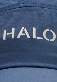 Niebieska czapka z teksturowanej tkaniny z białym logo "HALO" na przodzie. Projekt ma strukturalny kształt z zakrzywionym daszkiem.