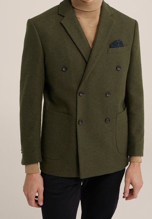 Homme portant un blazer croisé vert olive avec des boutons noirs, un pull à col roulé beige, un pantalon noir et un mouchoir de poche bleu marine à motifs.