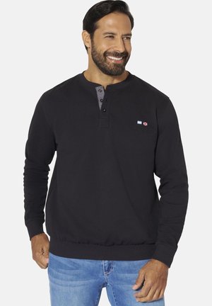 Smiling man with dark hair and beard wears a black long-sleeve henley shirt and light blue jeans, standing against a white background.