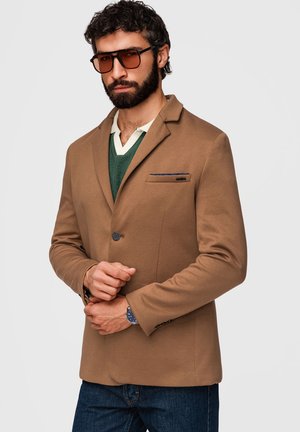 Mand med skæg og krøllet hår iført en brun blazer, grøn sweater, hvid skjorte, solbriller og blå jeans, der justerer sit ur.