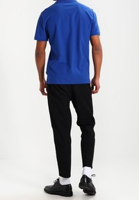 Man i en blå polo-tröja, svarta byxor, vita strumpor och svarta fin-skor som står med ryggen mot kameran på en vit bakgrund.