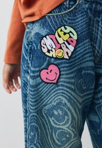 Jeans din denim cu un model abstract albastru, având broderii colorate: "LUMEA ZÂMBETELOR" sub formă de inimă și o inimă roz cu un față zâmbitoare.