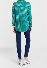 Blouse verte à pois avec manches longues, col boutonné et ourlet arrondi, associée à un jean skinny foncé et des chaussures à talons blocs blanches.