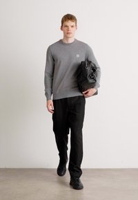 Grauer Crewneck-Pullover, schwarze Hose und schwarze Sneakers; das Model hält eine schwarze Tasche mit strukturierter Oberfläche und geht vor einem neutralen Hintergrund.