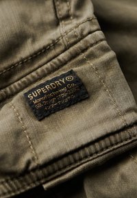 Olive green fabric with a soft texture, featuring a black label with gold text. The label reads "SUPERDRY" and includes care instructions.