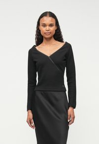 Top preto com nervuras, de mangas compridas e decote em V, com um design em drapeado e um pequeno logotipo bordado perto da bainha, combinado com uma saia de cetim.