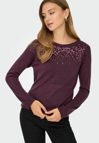 Pull violet avec des embellissements en sequins au niveau de l'encolure. Manches longues, texture douce et design ajusté. Accompagné de pantalons noirs.