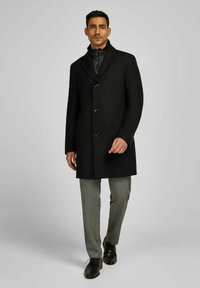 Cappotto nero in misto lana con design aderente, dotato di tre bottoni, colletto a scopertura e tasche laterali, abbinato a pantaloni grigi su misura.