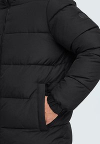 Svart pufferjakke med quiltet design, stående krage, elastiske mansjetter og håndlommer. Glatt stoff med rundt logo detaj på ermet.