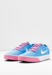 Blå kanvassneakers med rosa snören och en rosa sula; har en vit Nike-logga och en låg profildesign. Slät textur över hela.