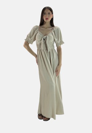 Robe longue beige avec des manches bouffantes et un détail de nœud à l'avant. Le corsage présente un design smocké, descendant vers une jupe fluide. Portée avec des sandales noires.