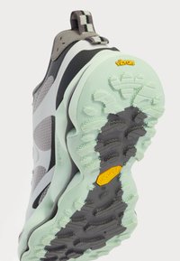 Chaussure de sport avec une tige texturée grise et noire, dotée d'une semelle en Vibram vert menthe. Les détails notables incluent un motif en rayures et une languette.