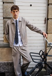 Młody mężczyzna w beżowym garniturze i krawacie opierający się o czarny rower przed białymi drewnianymi drzwiami i kamienną ścianą.