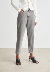 Mujer con un top blanco ajustado de manga larga que lleva unos pantalones vaqueros grises de pierna ancha y de cintura alta, además de unas sandalias de tacón negro y abierto, contra un fondo liso.